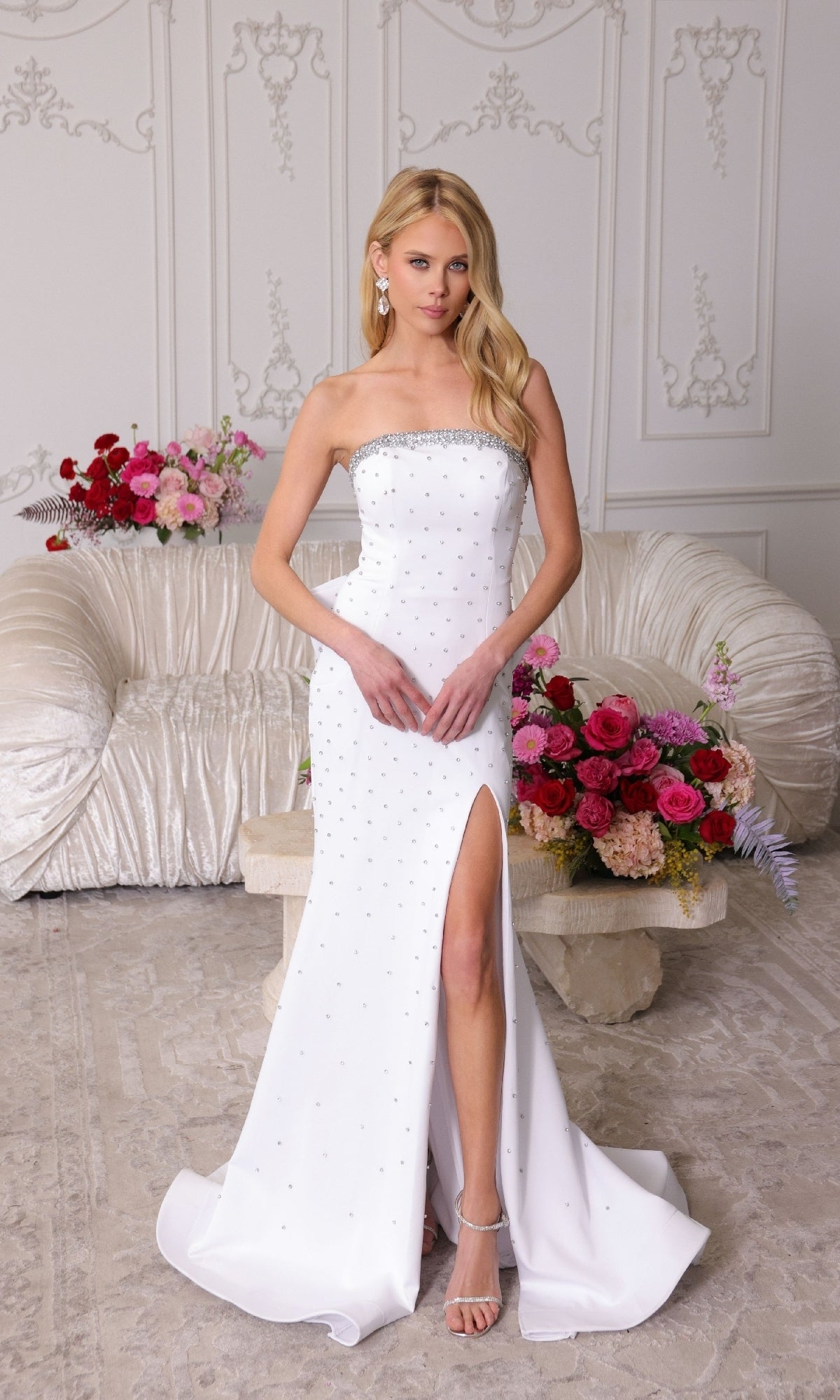 A woman wears the Strapless Long Bow Prom Dress by Ava Presley 40153, with rhinestone accents and a thigh-high slit, standing indoors amid floral arrangements and an ornate white sofa.